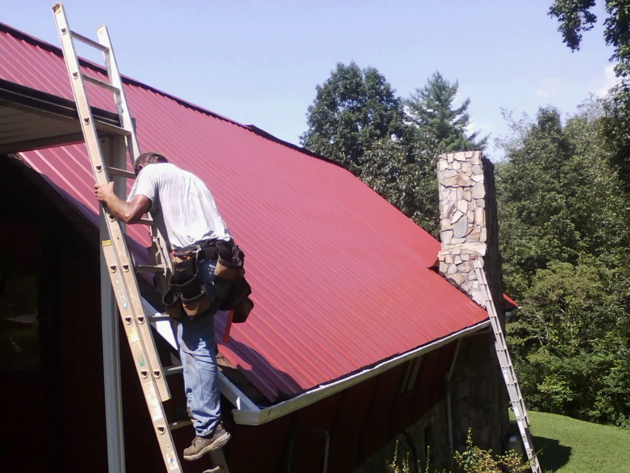 Roofer on a ladder beside a red metal roof and stone chimney — Soffit & Fascia Repair by Roofing Brothers in Trout Run